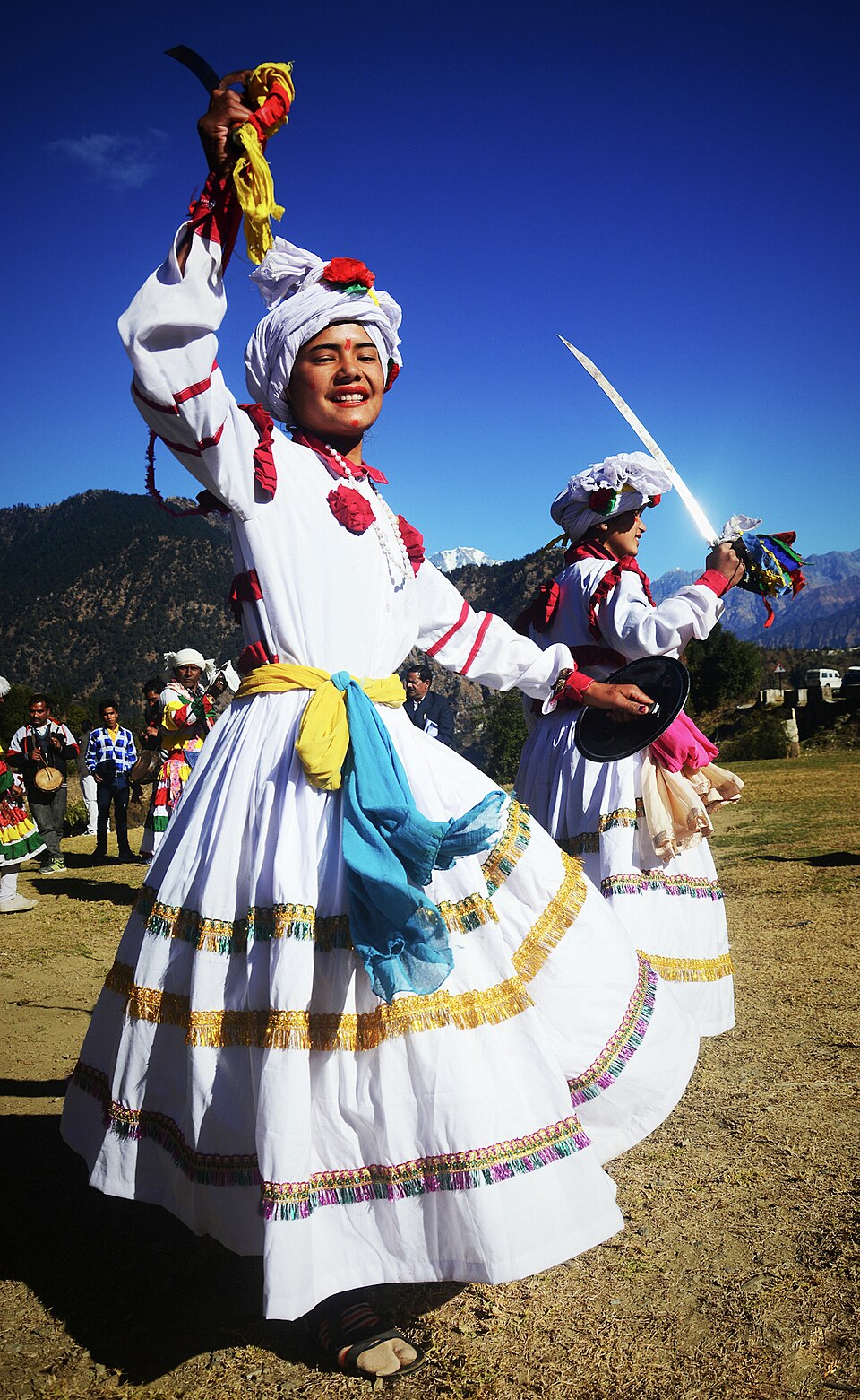 A chholiya dancer in white robes and turban with sword raised and brass shield, Himalayan peaks behind
